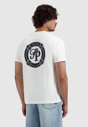 Man met krullend haar die een wit T-shirt draagt met een zwart rond "Pure Path"-logo en lichtblauwe spijkerbroek, staand opzij.