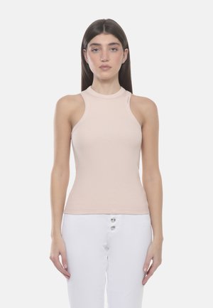 Giovane donna con capelli castani lisci che indossa un top beige a coste senza maniche e pantaloni bianchi con dettagli a bottoni, in piedi contro uno sfondo semplice.