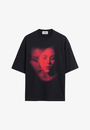 Homme aux cheveux courts portant un t-shirt noir avec un motif graphique rouge représentant le visage d'une femme en mouvement, assorti à un pantalon blanc, debout devant un fond uni.