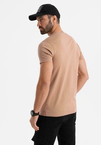 Béžové tričko s krátkým rukávem a kulatým výstřihem, spárované s černými cargo kalhotami. Outfit doplňuje černá kšiltovka a na zápěstí je nasazené hodinky.