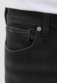 Detail vrecka čiernych rifle s viditeľným prešitím, kovovým nitom a pútkom na opasok, nosené s bielou košeľou.