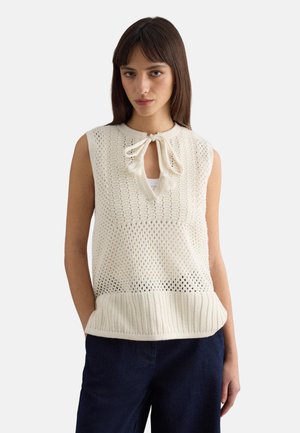 Donna con top senza maniche color crema lavorato a maglia, con chiusura a fiocco sul collo, e pantaloni blu navy, in piedi davanti a uno sfondo semplice.