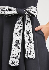Abito nero con una cintura in pizzo bianco annodata a fiocco, che mostra motivi floreali nel pizzo, realizzato in tessuto liscio e un design moderno.
