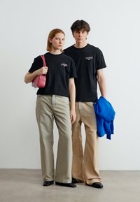 Νεαρή γυναίκα και άνδρας φοράνε μαύρα μπλουζάκια με σχέδια και μπεζ παντελόνια με φαρδιά μπατζάκια· η γυναίκα κρατάει κόκκινη τσάντα, ο άνδρας κρατάει μπλε ζακέτα.