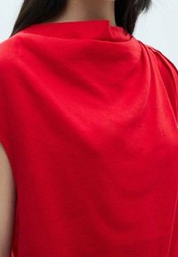 Rotes ärmelloses Oberteil mit einem drapierten Ausschnitt und glatter Textur. Der Stoff wirkt leicht, mit klaren Linien und minimalistischem Design.