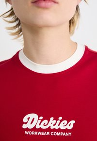 Raudona medvilninė marškinė su balta apykakle. Priekyje ant krūtinės puikuojasi didelis baltas logotipas "Dickies WORKWEAR COMPANY".