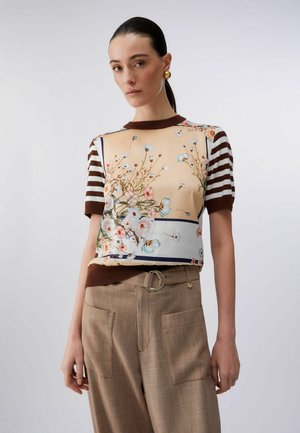 Donna che indossa una maglietta a maniche corte con stampa floreale, maniche a righe marroni e pantaloni beige a vita alta con grandi tasche e cintura.