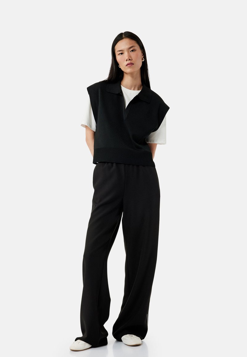 Gilets en tricot noir avec un col sur un t-shirt blanc, associé à un pantalon noir ample et des chaussures blanches à enfiler. Design simple et décontracté.