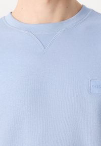 Felpa azzurra con tessuto strutturato, collo a girocotolo a coste e un piccolo patch logo rettangolare sul petto. I dettagli delle cuciture sono visibili.
