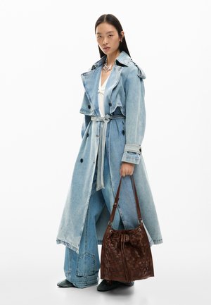 Vrouw in een lange lichtblauwe denim trenchcoat en wijduitlopende jeans, met een grote bruine geweven leren handtas, tegen een witte achtergrond.