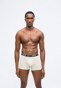 Μυώδης άνδρας που φοράει ανοιχτό γκρι μποξέρ Nike με μαύρη ζώνη μέσης, στέκεται μπροστά σε απλό φωτεινό φόντο, κοιτάζοντας μπροστά.