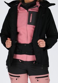 Black outer jacket with zippers, pink fleece inner layer, contrasting fabric accents, and adjustable waistband. Features functional pockets.