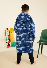 Blauer Camouflage-Hoodie mit lockerer Passform und weicher Textur, kombiniert mit schwarzen Hosen. Im Hintergrund sind Skateboards und ein grüner Stuhl.