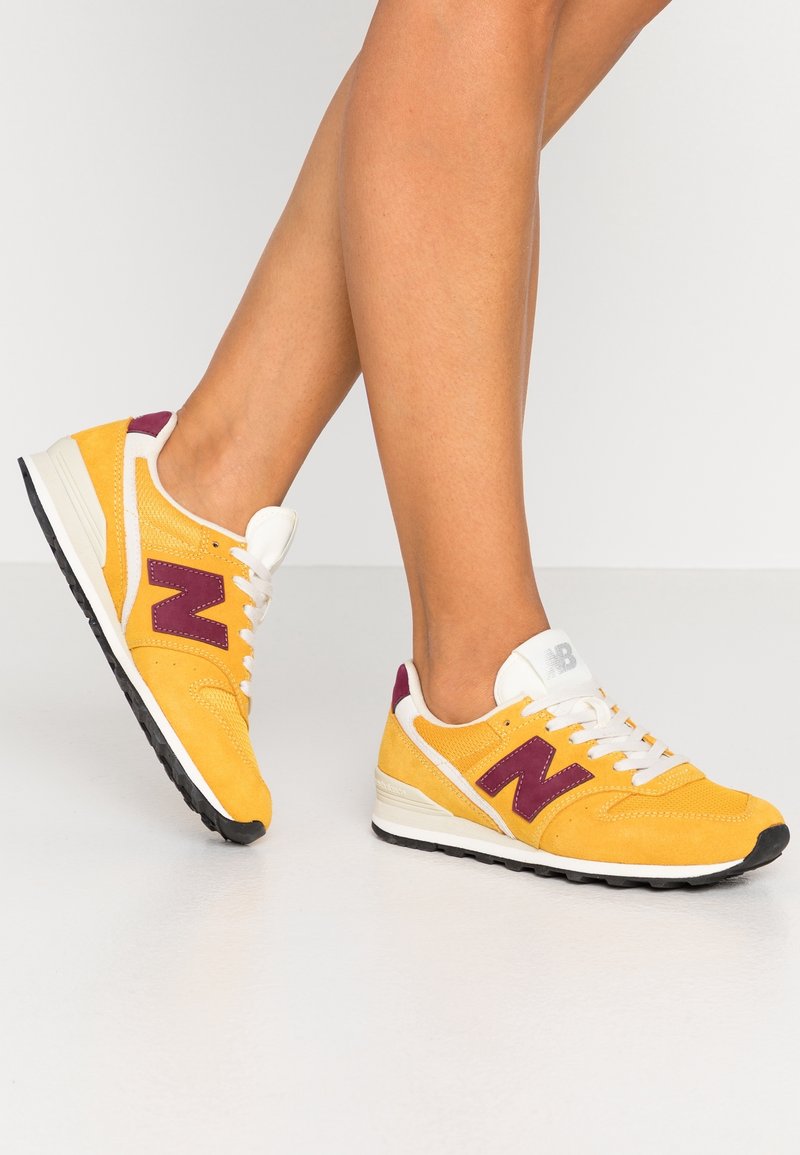 Ben klädda i gula och lila New Balance-sneakers med vita skosnören, stående på en glänsande vit golvyta.