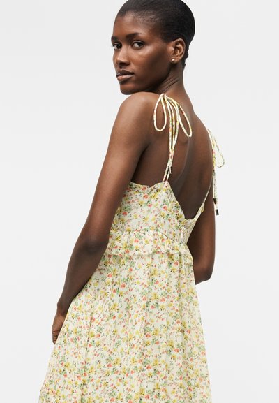 Γυναίκα με σκούρο δέρμα και κοντά μαλλιά, που φοράει ένα λουλουδάτο φόρεμα με λεπτές τιράντες και ανοιχτή πλάτη, κοιτάζοντας πάνω από τον ώμο της.