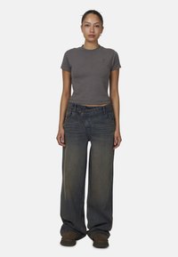 T-shirt gris cropped et jeans en denim bleu à jambe large. Le T-shirt a un petit logo ; les jeans sont amples avec une finition délavée. Chaussures brunes.