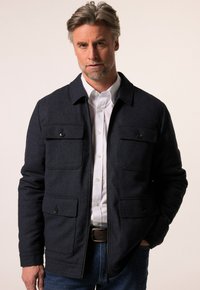 Tmavě modrá bunda vyrobená z texturované látky s límcovým designem, čtyřmi předními kapsami a zapínáním na zip, doplněná bílou košilí.