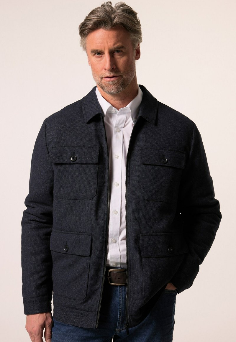 Tmavě modrá bunda vyrobená z texturované látky s límcovým designem, čtyřmi předními kapsami a zapínáním na zip, doplněná bílou košilí.