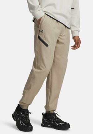 Beige jogger pants with black zippered pocket, worn with white long-sleeve top and black athletic shoes with white accents.
