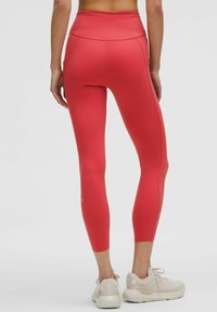 Czerwone legginsy z wysokim stanem i dopasowanym krojem. Zauważalne detale szwów oraz boczna kieszeń. W połączeniu z jasnoszarymi butami sportowymi.