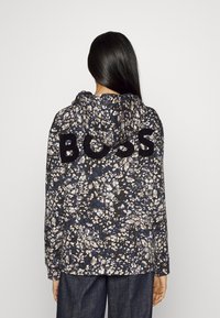 Sudadera con capucha en un patrón floral oscuro con tonos de azul, negro y beige; "BOSS" en grandes letras negras en la parte posterior.
