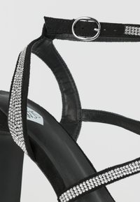 Sandal con tacco alto nero con cinturini sottili, decorato con accenti di strass lungo i bordi. Realizzato in camoscio con una finitura liscia. Dettaglio con fibbia metallica.