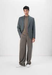 Grå blazer över en brun polotröja, kombinerad med lösa, ljusgrå byxor och vita sneakers. Snyggt tyg med skräddarsydd design.