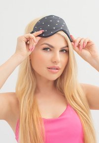 Mascherina nera per il sonno con pois bianchi, leggermente imbottita, tenuta da una persona con lunghi capelli biondi. Tessuto liscio, morbido al tatto.