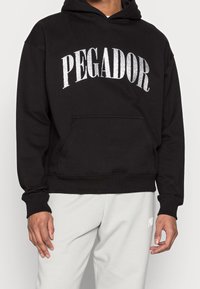 Felpa nera con la scritta "PEGADOR" in lettere di paillettes argento. Presenta una tasca frontale e polsini e orlo a costine. Indossata con pantaloni da jogging grigi chiari.