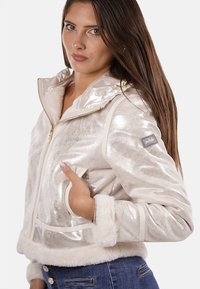 Giacca argentata metallica con colletto e polsini foderati in pelliccia, caratterizzata da una chiusura frontale con zip, tasche laterali e un logo sul maniche.