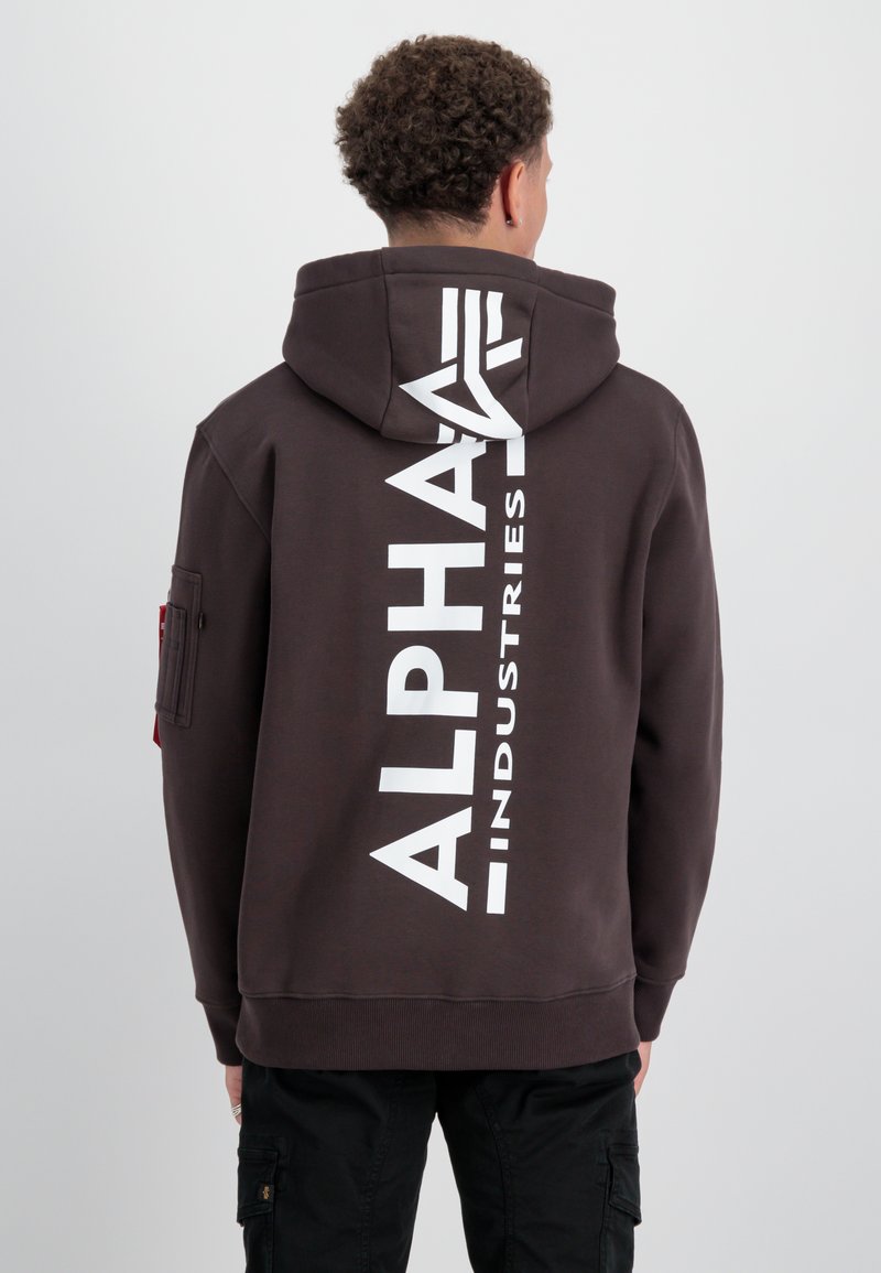 Sudadera marrón con un gran logo blanco que dice "ALPHA INDUSTRIES" en la parte de atrás, con capucha ajustable y un bolsillo lateral.