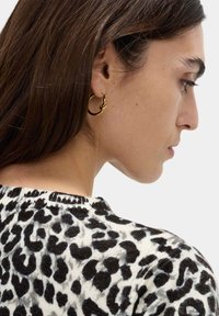 Guldörhänge i form av en snodd design, bärs av en person med långt mörkt hår; i bakgrunden syns en svartvit leopardmönstrad tröja.