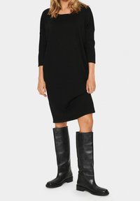 Abito nero oversize con maniche a tre quarti, realizzato in tessuto morbido. Abbinato a stivali neri in pelle alti fino al ginocchio con punta rotonda.