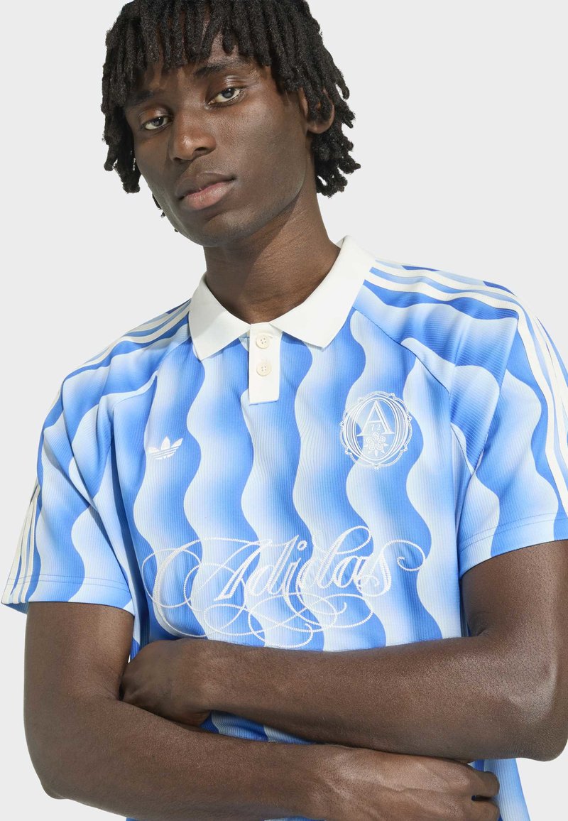 Junger Mann mit dunkler Haut und Dreadlocks, der ein blau-weiß gestreiftes Adidas Polo mit wellenförmigen Streifen trägt, Arme verschränkt, neutraler Gesichtsausdruck.