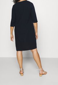 Mörkblå, knälång klänning med trekvartslång ärm, slät textur och avslappnad passform. Matchas med ljusa, remmade sandaler.