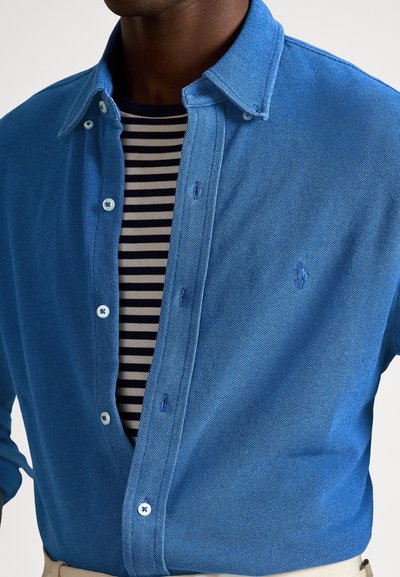 Mėlyna sagomis užsegama marškinėliai iš tekstūruotos medžiagos, su plėčiamu apykakliu ir atitinkančiomis sagomis. Dėvimi ant juodai baltų dryžuotų marškinių.