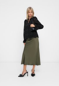 Femme portant un haut en dentelle noire, une jupe midi satin olive, des chaussures à talons hauts noires et tenant un petit sac à bandoulière noir.
