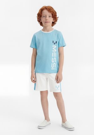 Niño pelirrojo de pie mirando hacia adelante, vestido con camiseta azul claro de Messi y pantalones cortos blancos con el logo de Messi, zapatos blancos sin cordones.