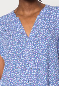 Kvinde iført en blå bluse med et lille pink og hvidt blomsterprint og V-udskæring.