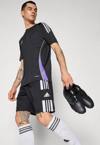 Svart atletisk skjorte med lilla og grønne detaljer, kombinert med svarte shorts med hvite striper og Adidas-logo. Svarte fotballsko i hånden.