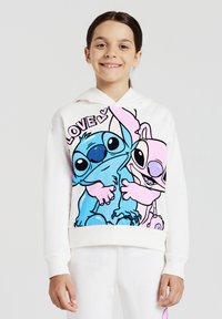 Ragazza sorridente che indossa un hoodie bianco con i colorati personaggi Stitch e Angel che si abbracciano, con la parola "AMORE" e cuori sopra.