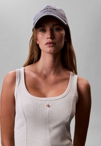 Νέα γυναίκα με μακριά μαλλιά που φοράει ανοιχτό γκρι καπέλο Calvin Klein και λευκή ριμπ τοπ μπλούζα με το λογότυπο CK στο στήθος, πάνω σε απλό γκρι φόντο.