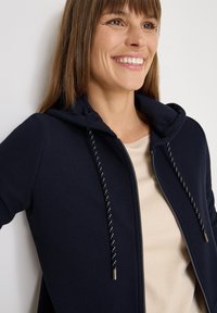 Donna sorridente che indossa una camicia beige e una felpa con zip blu navy con cappuccio e cordini, in piedi su uno sfondo bianco.