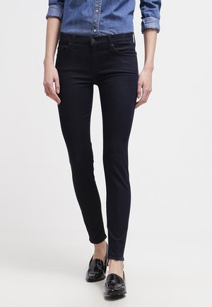Kvinde iført mørke skinny jeans, sorte loafers og en blå denim skjorte med opkrogede ærmer, stående på en ensfarvet baggrund.