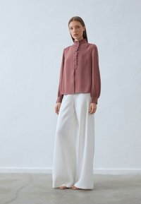 Blusa bordeaux con parte anteriore a volants, colletto alto, maniche lunghe e chiusura con bottoni, abbinata a pantaloni bianchi a gamba larga a vita alta.