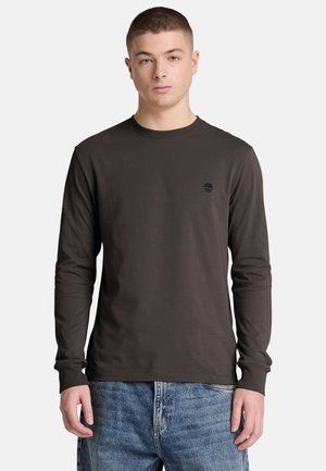 Jonge man die een donker lang mouwen shirt en blauwe spijkerbroek draagt, staat voor een effen lichte achtergrond.