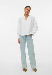 Camicia bianca con scollo a V, maniche lunghe e polsini elastici, abbinata a jeans azzurri a vita alta e scarpe basse color taupe.
