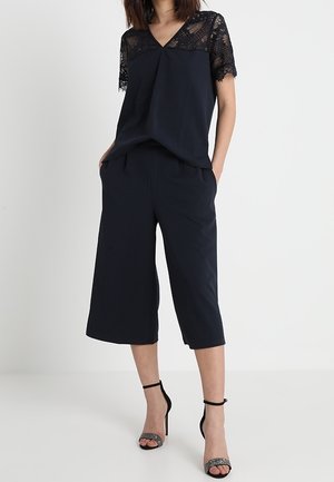Femme portant un haut en dentelle noir à manches courtes, un pantalon noir ample et court, et des sandales à talons hauts avec bride à la cheville, debout devant un fond blanc.