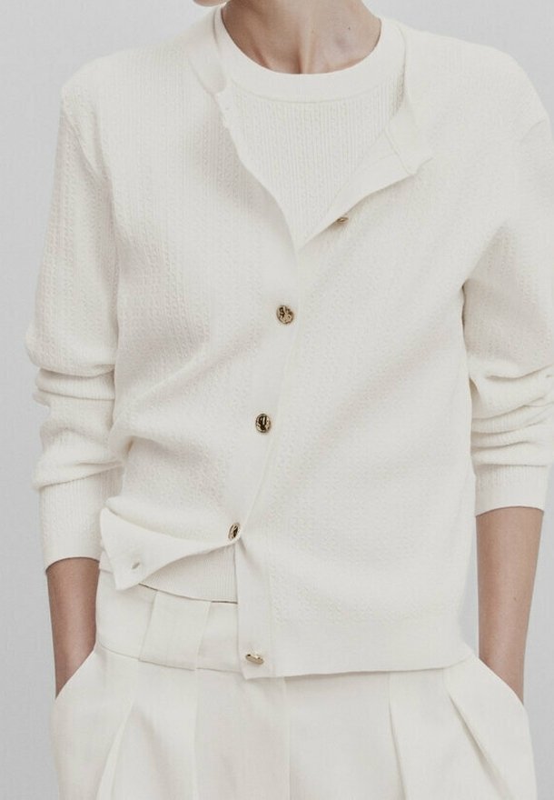 Personne portant un cardigan en tricot couleur crème avec des boutons, par-dessus un pull assorti et un pantalon plissé taille haute, mains dans les poches.