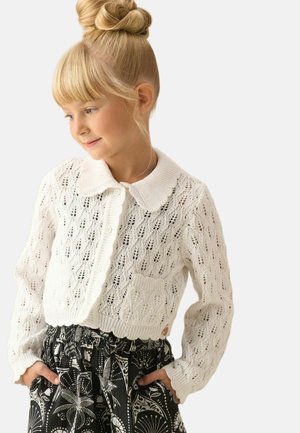 Jeune fille aux cheveux blonds en chignon, portant un cardigan blanc en maille et un pantalon noir à motifs, regardant vers le bas avec les mains dans les poches.